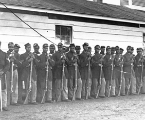 United States Colored Troops soldiers standing with rifles in front of wooden building.