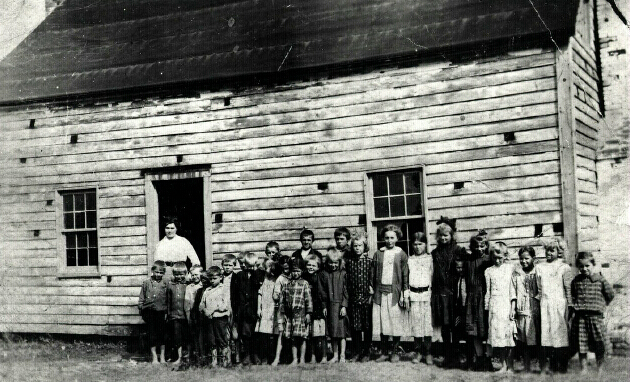 students and teacher in row outside schoolhouse